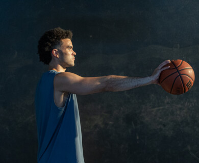 A person is holding a basketball with an extended arm in a dimly lit environment, wearing a sleeveless shirt. Person holding a basketball with an extended arm, wearing a sleeveless shirt in dim light.