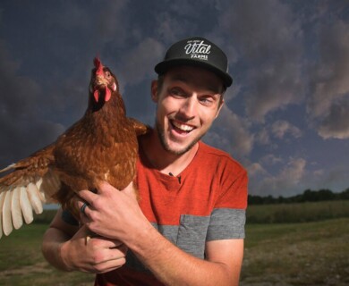 A person wearing a cap and a red shirt is holding a brown chicken outdoors, with a cloudy sky and grassy field in the background. Person in cap and red shirt holding a brown chicken outdoors with cloudy sky and grassy field.