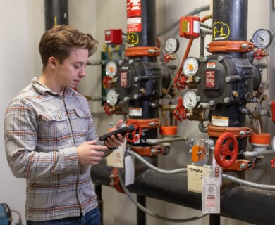 A person in a plaid shirt is inspecting machinery with pipes and gauges, using a handheld device in an industrial setting. Person in plaid shirt inspects machinery with handheld device in an industrial setting.