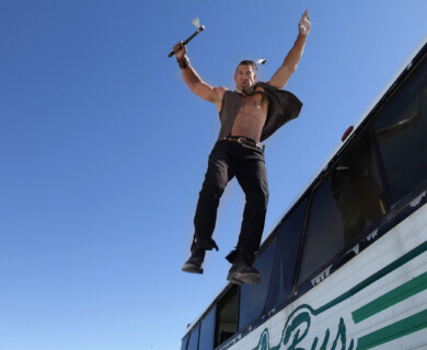 A person is jumping from the roof of a bus, holding a small axe in one hand. The sky is clear and blue in the background. Person jumping from bus roof holding small axe, clear blue sky background.