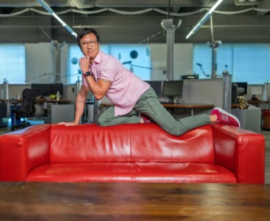 A person is posed kneeling on a vibrant red couch in an office environment. The individual is wearing a pink shirt, green pants, and maroon sneakers. Office furniture and computers are visible in the background, with overhead lighting and windows adding to the setting. Person kneeling on red couch in office; pink shirt, green pants, maroon sneakers visible.