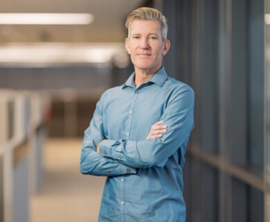 A person is standing in an office environment, wearing a light blue button-up shirt, with arms crossed. The background shows a corridor with cubicles, and the lighting is bright and professional. Person in light blue shirt standing in bright office with cubicles and crossed arms.