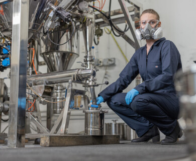 A person wearing a protective suit, gloves, and a respirator is kneeling beside industrial machinery in a laboratory or manufacturing setting. Person in protective gear kneels by industrial machinery in a lab or manufacturing area.