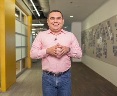 A person is standing in a hallway, wearing a pink button-up shirt and jeans. The individual is smiling and has a hands clasped in front. The hallway has bright lighting, with office elements like windows and bulletin boards visible in the background. Person in pink shirt and jeans smiling in office hallway with bulletin boards and windows.