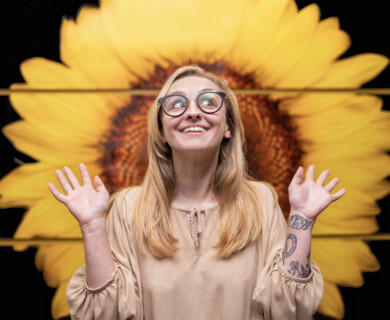 A person with long hair and glasses is smiling broadly while standing in front of a large, vibrant sunflower backdrop. The individual has tattoos on their forearm and is wearing a light-colored top, raising their hands in an expressive gesture. Person with long hair and glasses smiling in front of a vibrant sunflower backdrop, arms raised.