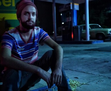 A person wearing a striped shirt and a head covering is sitting on a curb at a dimly lit gas station. In the background, there is a vintage car parked near the gas pumps. The scene is set at night, with a subdued lighting ambiance. Person in striped shirt and head covering sitting at night gas station near vintage car.