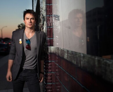 A person is standing outdoors at sunset, leaning against a brick wall with a reflective surface. They are wearing a gray blazer, gray shirt, and dark pants, with sunglasses hanging from their shirt. The reflection of the person is visible in the glossy surface of the wall. Person outdoors at sunset, leaning on brick wall with reflection, wearing gray attire and sunglasses