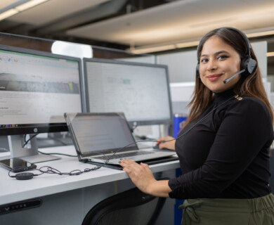 A person wearing a headset is standing at a desk with multiple computer monitors and a laptop. The office environment has modern decor, and there is a smartphone and audio device on the desk. Person wearing headset at desk with monitors, laptop, smartphone, in modern office setting.