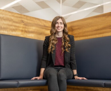 A person with long, wavy hair sits on a blue cushioned bench against a wooden wall, wearing a black blazer and a maroon top. Person with wavy hair in blazer and maroon top sits on blue bench by wooden wall.