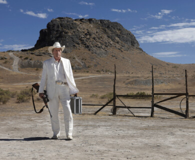 A person wearing a white suit and cowboy hat is walking in a desolate landscape. They are holding a briefcase and a rifle, with a rocky hill and a partially broken wooden fence in the background. The sky is clear and blue. Person in white suit holds rifle and briefcase, standing in desolate landscape, rocky hill background.