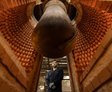 A person in protective clothing stands inside a large, cylindrical brick structure, possibly a kiln or industrial furnace, with a pipe or large object suspended above. The scene suggests a work environment related to manufacturing or heavy industry. Person in protective gear inside a brick kiln with pipe overhead, suggesting industrial work.
