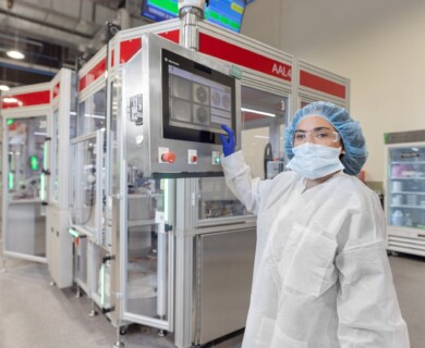A person in protective clothing, including a mask, gloves, and hair covering, operates a touchscreen panel in a sterile manufacturing environment, surrounded by industrial equipment and machines. Person in protective gear using touchscreen in sterile manufacturing environment.