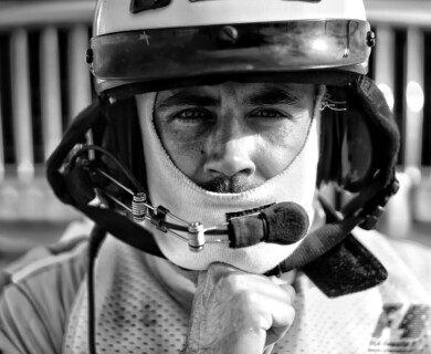 A grayscale image of a person wearing a racing helmet and a balaclava, with a microphone attached near the mouth. The background shows a blurred grille, possibly of a vehicle. Person in racing helmet and balaclava with microphone, blurred grille background.