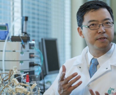 A person wearing a lab coat and glasses is speaking, gesturing with their hands in a scientific laboratory setting. There is laboratory equipment and instruments in the background. Person in lab coat and glasses gesturing in a laboratory with equipment in background.