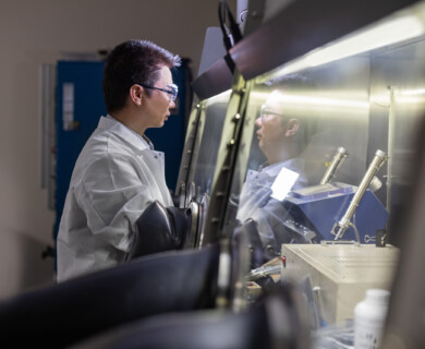 A scientist wearing safety glasses and a lab coat works inside a laboratory glove box, handling materials or conducting an experiment in a controlled environment. Scientist in safety gear working in a laboratory glove box handling materials.
