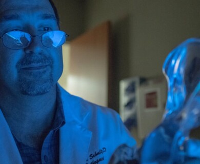 A man wearing glasses and a lab coat is observing a glowing blue object in a dimly lit room. Man in glasses and lab coat observing glowing blue object in dimly lit room.
