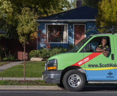 A person is smiling while leaning out of the driver's window of a colorful van parked on a suburban street. The van has a green and blue design with information about a business, including a website and a "100% satisfaction guarantee." A small house with a blue exterior and a red door is in the background, surrounded by greenery and trees. Person smiling from a colorful business van parked in front of a blue house.