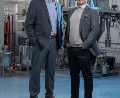Two individuals in formal attire stand in an industrial setting, featuring large metal equipment and machinery in the background. Two individuals in suits stand in an industrial area with large metal machinery behind them.
