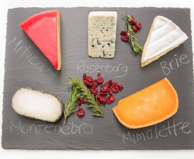 A slate platter features various cheeses labeled as Mitica, Rosenborg, Brie, Montenebro, and Mimolette. The cheeses are arranged alongside sprigs of rosemary and dried cranberries for decoration. Mitica, Rosenborg, Brie, Montenebro, Mimolette.