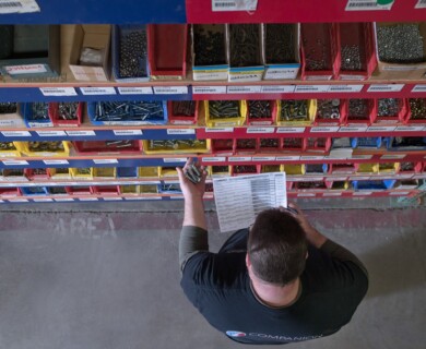A person is holding bolts in one hand and a checklist in the other while standing in front of shelves filled with labeled bins of various small hardware items, such as screws and bolts, in a storage or warehouse setting. Person holding bolts and checklist in front of bins with screws and bolts in a warehouse.