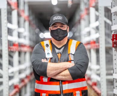 A person wearing an orange and white safety vest, protective face mask, and a cap is standing with arms crossed in a warehouse aisle. Person in orange safety vest, mask, and cap stands with arms crossed in a warehouse aisle.