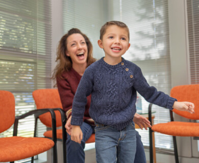 A woman and a young boy are in a room with orange chairs and blinds on the windows. The boy is smiling and wearing a blue sweater, while the woman, seated behind him, is laughing. Woman and boy in room with orange chairs, blinds; boy smiling, wearing blue sweater.