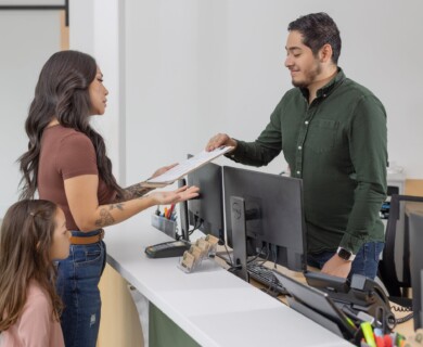 A woman and a child are standing at a reception desk. The woman is handing a clipboard to a man behind the desk, who is wearing a green shirt. Several computer monitors and office supplies are visible on the counter. Woman and child at reception desk, woman handing clipboard to man in green shirt.