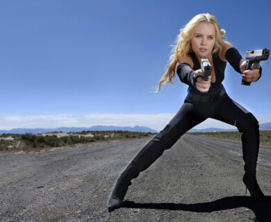A woman wearing black clothing is crouched on a deserted road, holding a gun in each hand. The background features a clear blue sky and distant mountains. Woman in black crouching on empty road, holding guns, mountains and blue sky in background.