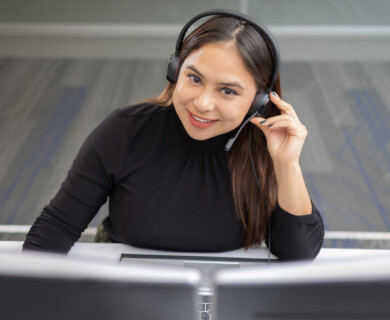 A woman wearing a headset is sitting at a desk with dual monitors. She is smiling and appears to be engaged in a conversation or call. The setting suggests an office or customer service environment. Woman in headset at desk with dual monitors, engaged in conversation, office setting.