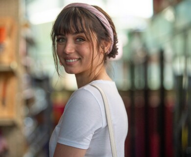 A woman with a pink headband and wearing a white t-shirt is smiling while turning slightly towards the camera. The background appears to be blurred shelves, suggesting an indoor setting. Woman with pink headband smiles, wearing a white t-shirt in a blurred indoor setting.