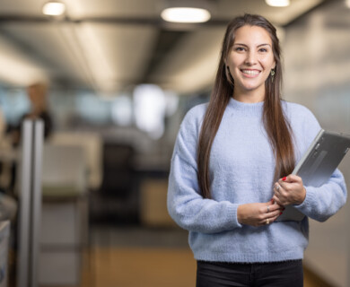 A woman in a light blue sweater stands in an office environment, smiling and holding a closed laptop. The background is blurred, with indistinct figures and office furnishings visible. Woman in light blue sweater holding a laptop, smiling in an office with blurred background.