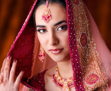 A woman wearing traditional attire adorned with intricate embroidery and beadwork. She has henna designs on her hand, and her jewelry includes earrings and a necklace, complemented by a headpiece. The background is softly blurred. Woman in traditional attire with embroidery, beadwork, henna, and jewelry; blurred background.