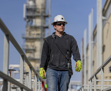 A person wearing a hard hat, safety glasses, and gloves is walking on a metal walkway at an industrial site. The background features a large structure, possibly part of the facility, under a clear blue sky. Worker in safety gear walking on industrial site metal walkway under clear sky.