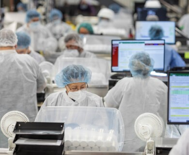 A group of workers in protective clothing, including masks and hairnets, are operating in a laboratory or manufacturing setting. They are surrounded by computer screens displaying data and are handling equipment or products in a controlled environment. Workers in protective gear operate computers in a lab or manufacturing environment.
