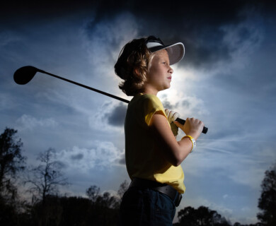 A young person wearing a visor and a yellow shirt is holding a golf club, standing against a dramatic sky with scattered clouds. They appear focused and prepared to play golf. Young golfer in yellow shirt and visor holding club under cloudy sky, appearing focused.