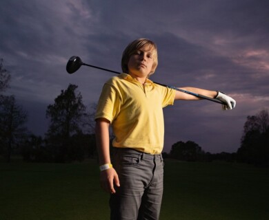 A young person wearing a yellow shirt and gray pants is standing outdoors against a cloudy sky, holding a golf club across their shoulders. They are wearing a white golf glove and a wristband on their right wrist, with trees visible in the background. Young person in yellow shirt outdoors, holding golf club on shoulders, cloudy sky, trees behind.