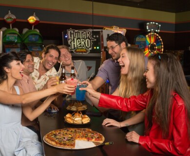 Group toasting with drinks at an arcade, pizza and sliders on table, Guitar Hero in background.