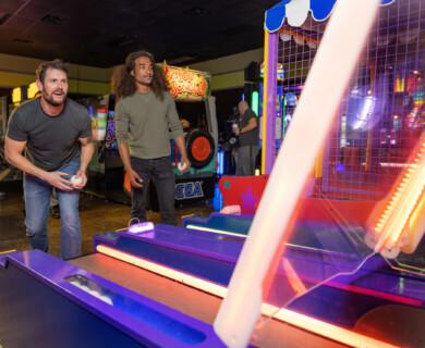 Two people are playing skee-ball in an arcade filled with colorful lights and various game machines. They appear focused on the game, holding balls and preparing to toss them. Two people playing skee-ball in an arcade with colorful lights and various game machines.