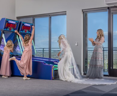 A bride in a white gown and veil is playing an arcade basketball game with two young girls wearing pink dresses. A woman in a light blue dress is clapping nearby. The setting appears to be indoors with large windows showing an outdoor view. Bride in gown plays arcade basketball with two girls, woman claps, indoor with outdoor view.