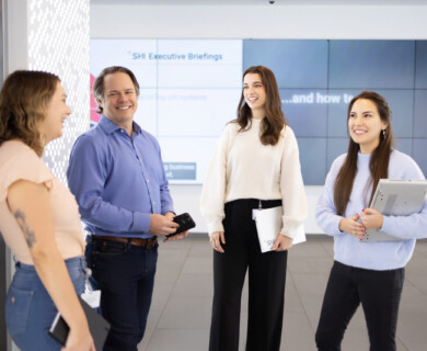 Four individuals in a professional setting, conversing with devices and folders.