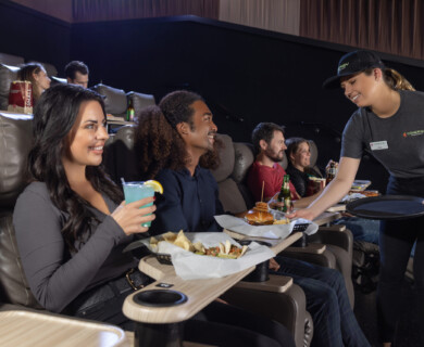 Cinema patrons in reclining seats enjoy food as a server delivers meals on trays.