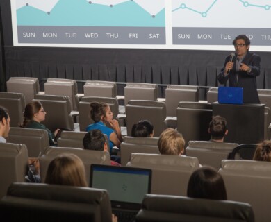Person presenting in a conference room with a graph on the screen and audience seated in rows.