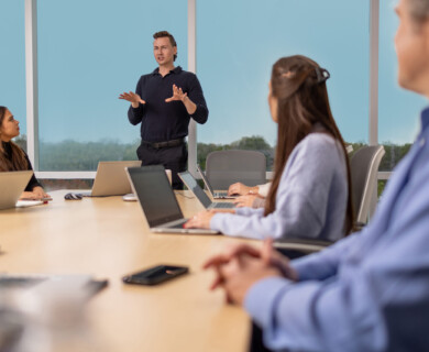 People in a conference room having a meeting, one standing, others seated with laptops, windows behind.