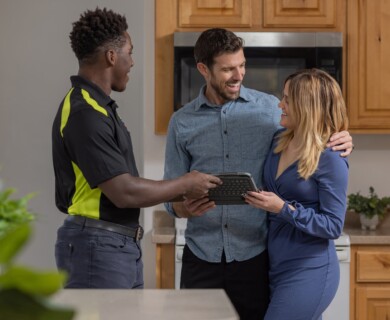 A couple is in a kitchen interacting with a person in a uniform holding a tablet. The atmosphere is friendly and the individuals appear engaged in conversation. Couple in kitchen talking with a uniformed person holding a tablet, friendly atmosphere.