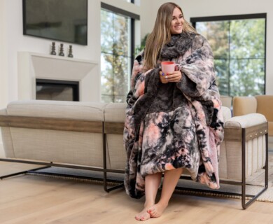 A person is sitting comfortably on a sofa, wrapped in a patterned, plush blanket. They are holding a cup, and the living room features large windows and modern decor, including a flat-screen TV mounted above a fireplace. Person cozy on sofa with patterned blanket, holding cup in modern living room with large windows.