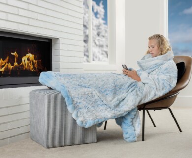 A person is sitting in a chair by a lit fireplace, wrapped in a fluffy blue blanket, and holding a smartphone. The room has a large window showing a snowy outdoor scene. Person wrapped in blue blanket, sitting by fireplace, using smartphone, snowy scene outside.