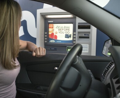 Person in a car using a drive-through ATM, screen shows message to provide info before leaving.