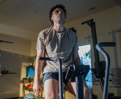 A person is sitting on an exercise bike in a gym setting, appearing focused. The gym contains various fitness equipment, and natural light comes through a window. Person focused on exercise bike in well-lit gym with various equipment.