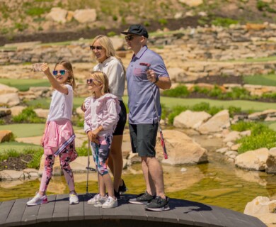 Family on a wooden bridge at mini-golf, children holding putters and taking a selfie.