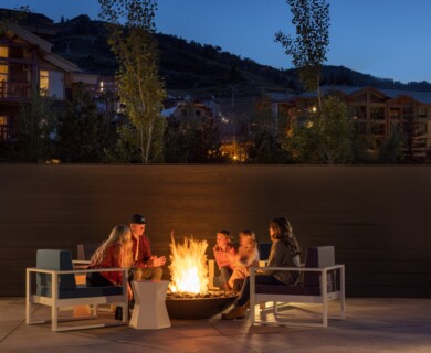 Group seated around fire pit with modern furniture, evening setting, hillside buildings visible.